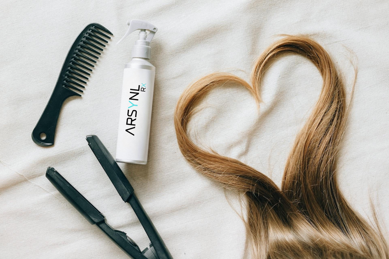 Hair care products including a bottle, comb, and straightener on a white surface with hair forming a heart shape.