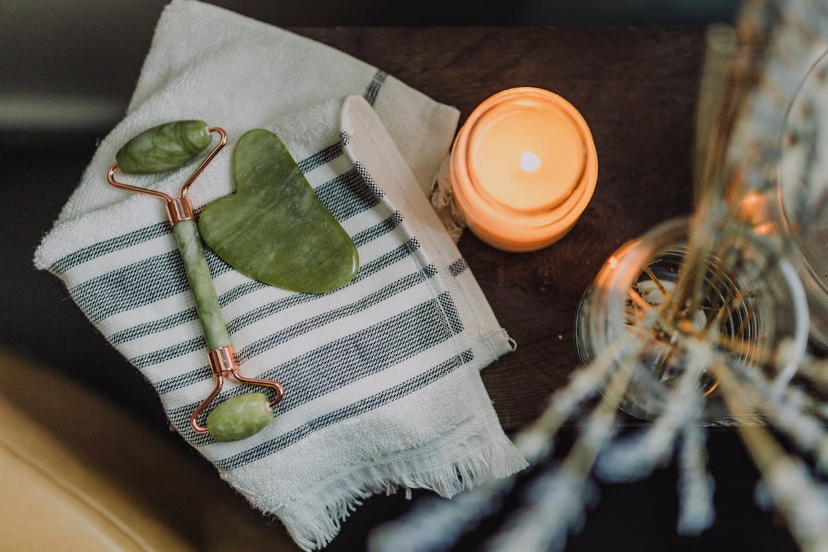 Skincare tools including a jade roller and face mask on a towel with a candle in the background.