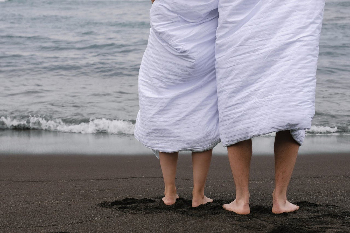 Man and woman standing on a beach wrapped in white towels while embraced lovingly.