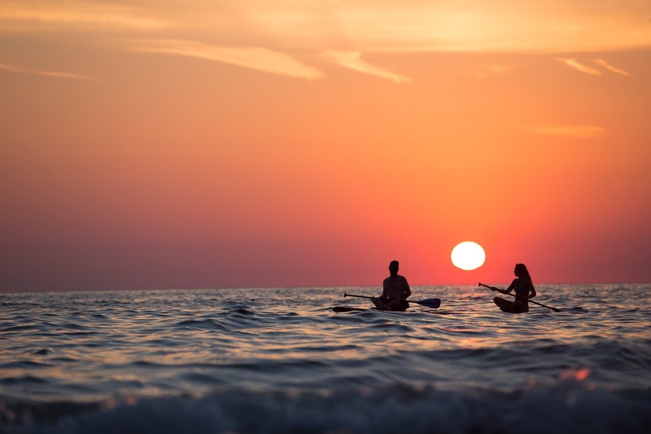 Healthy man and woman kayaking on the ocean at sunset with a large orange sun in the sky.
