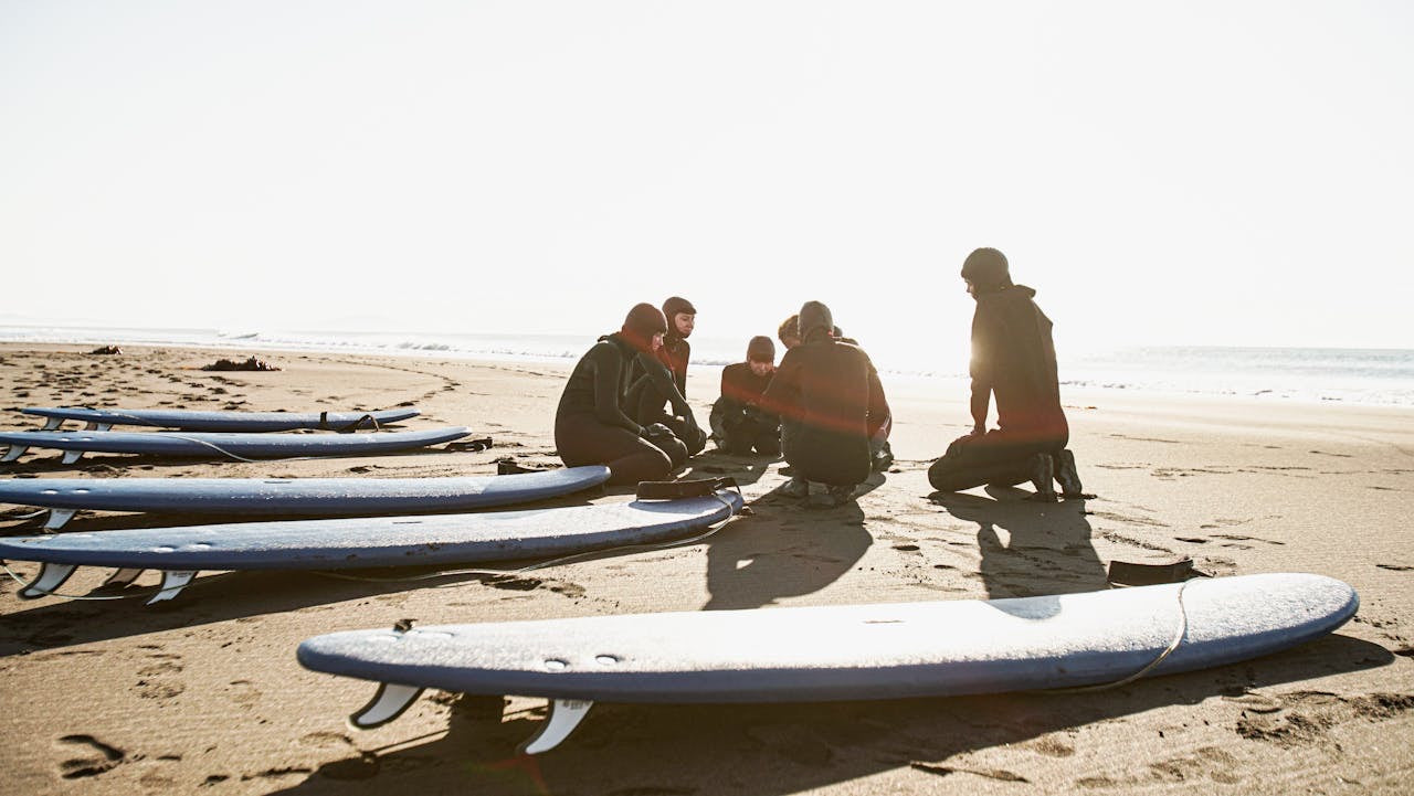 Group of surf athletes sitting on a sandy beach with surfboards lined up.